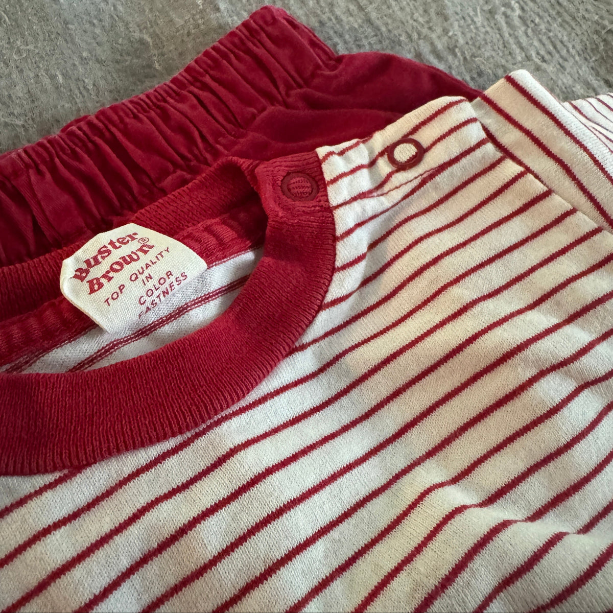 A close-up of a baby long-sleeve top by Peter Althouse, featuring a cream and red striped knit body with a solid red ribbed collar and elastic waistband.