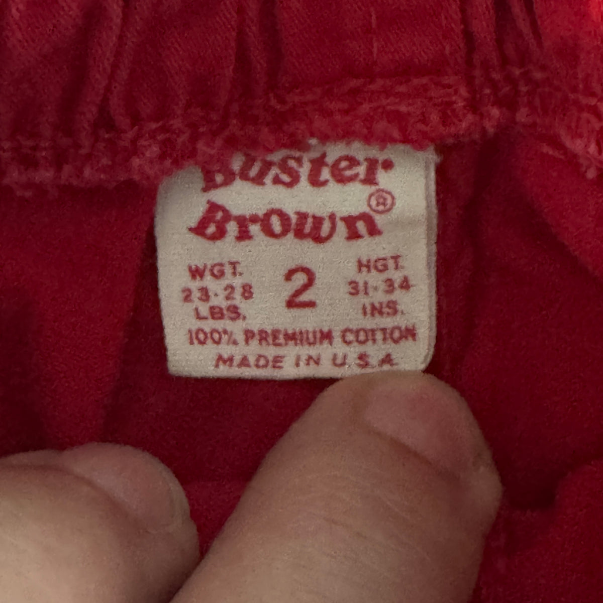 Close-up of a red garment's interior label reading 'Buster Brown' with size information, partially covered by a person's fingers.