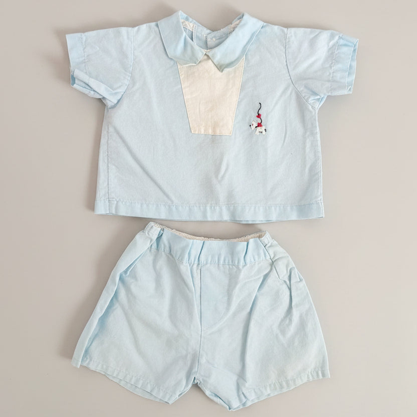 A baby boy's two-piece outfit consisting of a light blue short-sleeve collared shirt with a cream-colored chest panel and a small embroidered flamingo, paired with matching light blue shorts, laid flat on a textured brown fabric background.