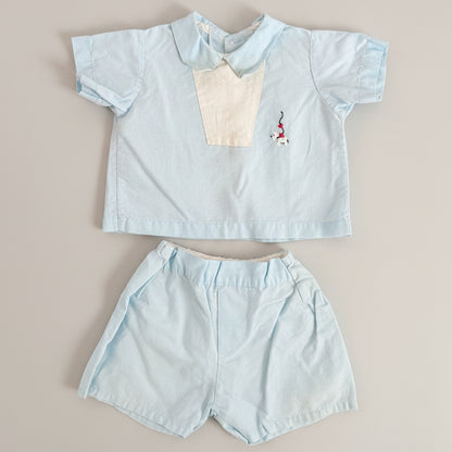A baby boy's two-piece outfit consisting of a light blue short-sleeve collared shirt with a cream-colored chest panel and a small embroidered flamingo, paired with matching light blue shorts, laid flat on a textured brown fabric background.