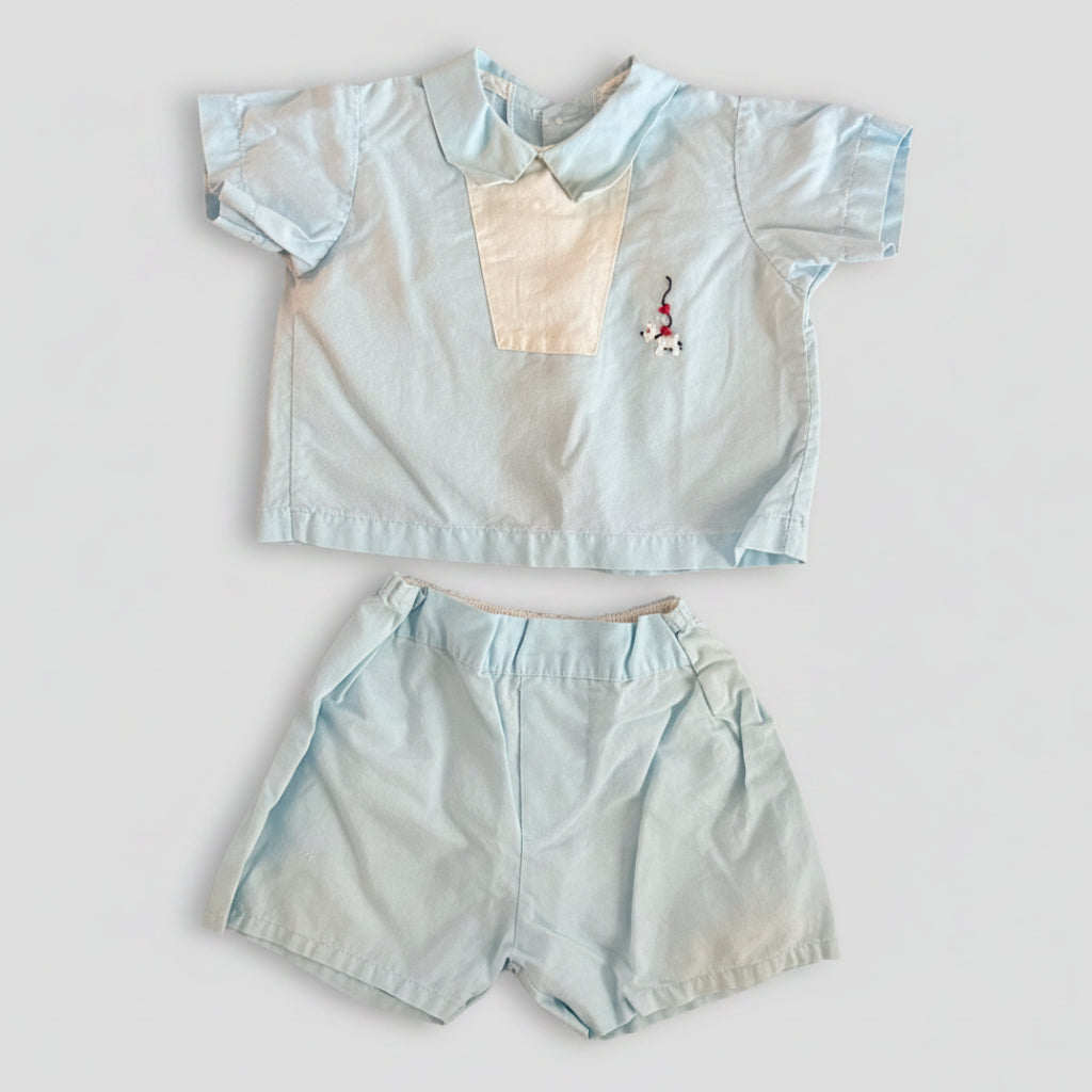 A light blue two-piece children's outfit set consisting of a short-sleeve collared shirt with a cream chest panel and a small embroidered character, paired with matching light blue shorts, laid flat on a textured brown fabric background.