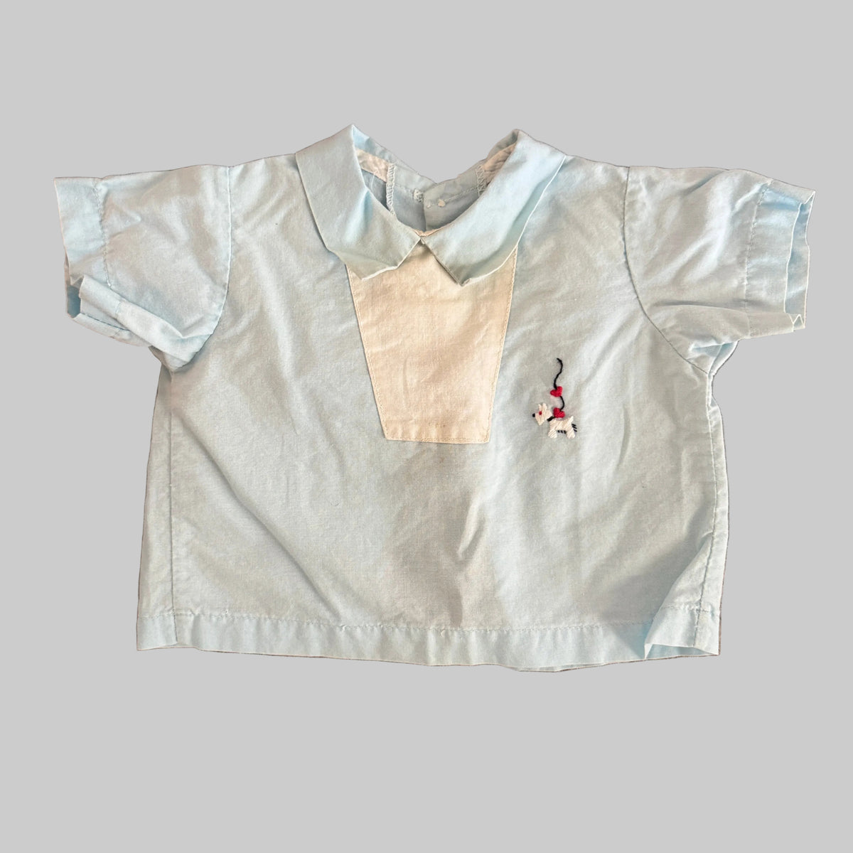 A light blue short-sleeve baby shirt with a cream-colored V-shaped chest panel and a small embroidered polo player logo on the left chest, laid flat on a brown textured fabric background.