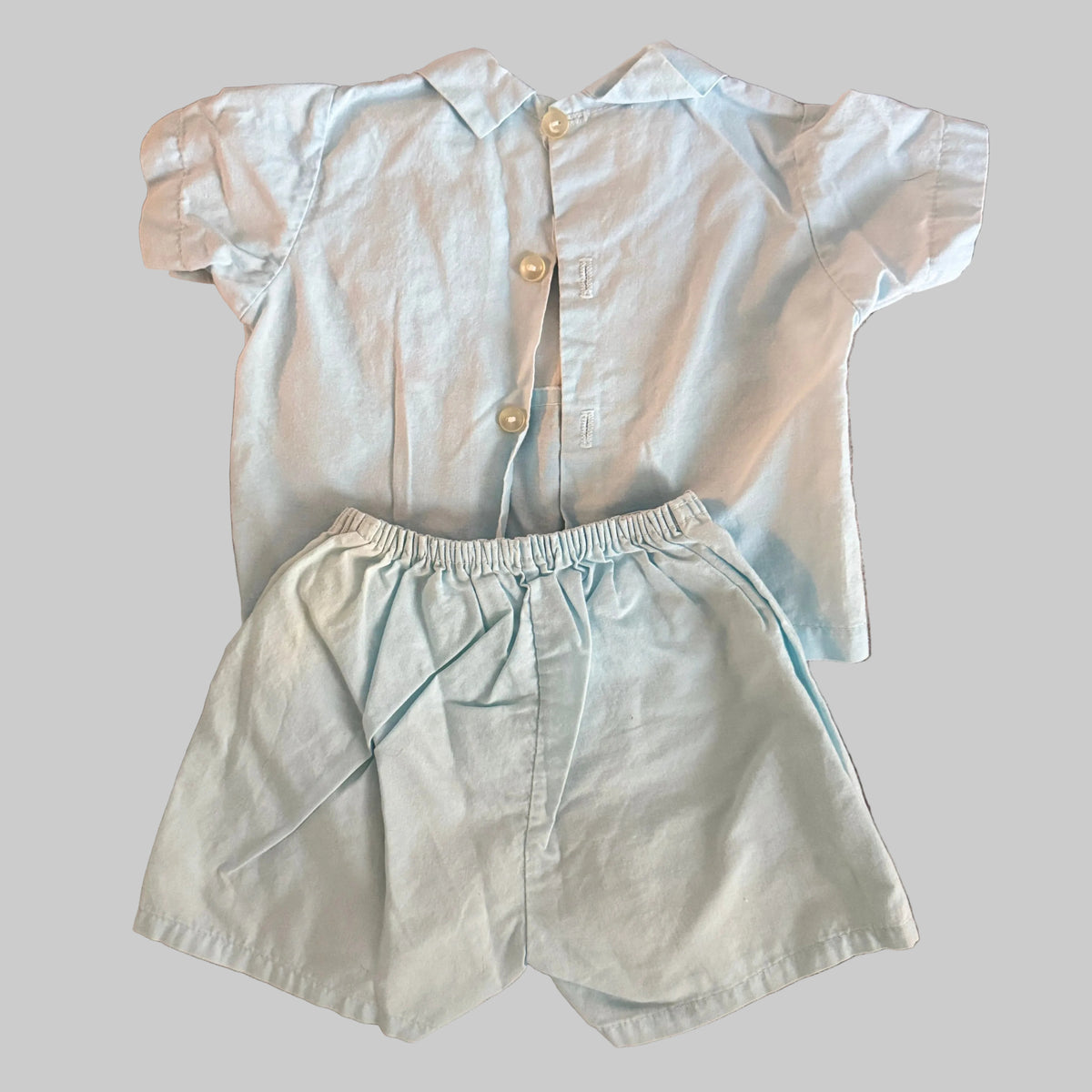 A baby boy's two-piece outfit consisting of a short-sleeve button-up shirt and matching shorts in a very light blue color, laid flat on a textured brown blanket.