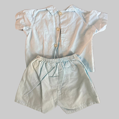 A baby boy's two-piece outfit consisting of a short-sleeve button-up shirt and matching shorts in a very light blue color, laid flat on a textured brown blanket.