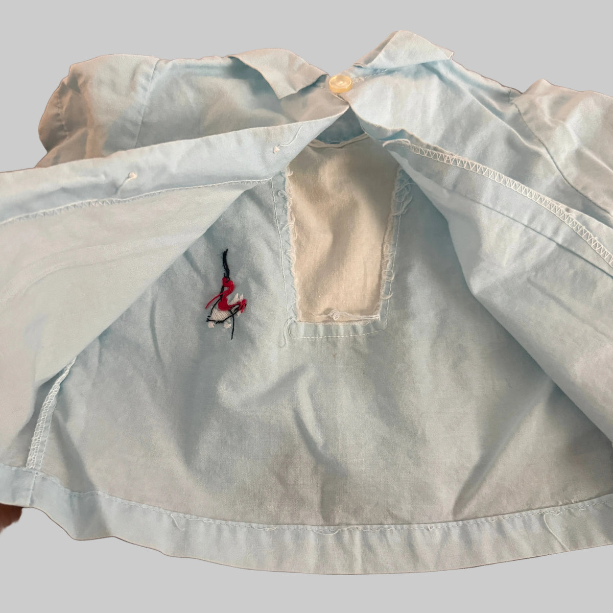 A light blue denim shirt laid flat on a textured brown fabric surface, showing a small embroidered logo on the chest and a cutout neckline.