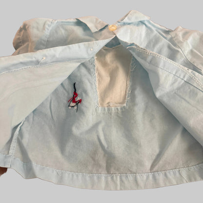 A light blue denim shirt laid flat on a textured brown fabric surface, showing a small embroidered logo on the chest and a cutout neckline.