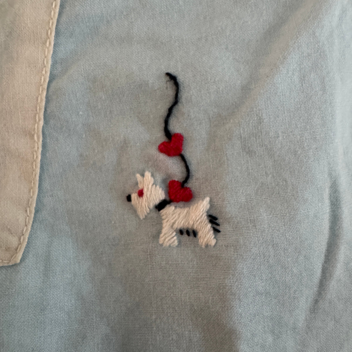 Close-up of a light blue fabric garment featuring an embroidered design of a white dog with red hearts and a black wavy line.