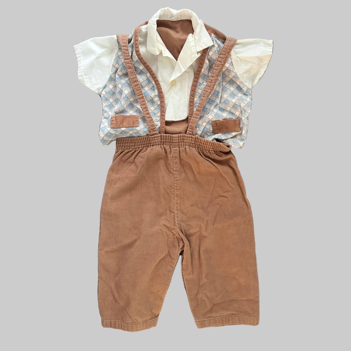 A baby boy's two-piece clothing set laid flat on a textured gray blanket. The set includes a brown corduroy bib-style overalls with suspenders and a white short-sleeve shirt with a light blue and white gingham vest.