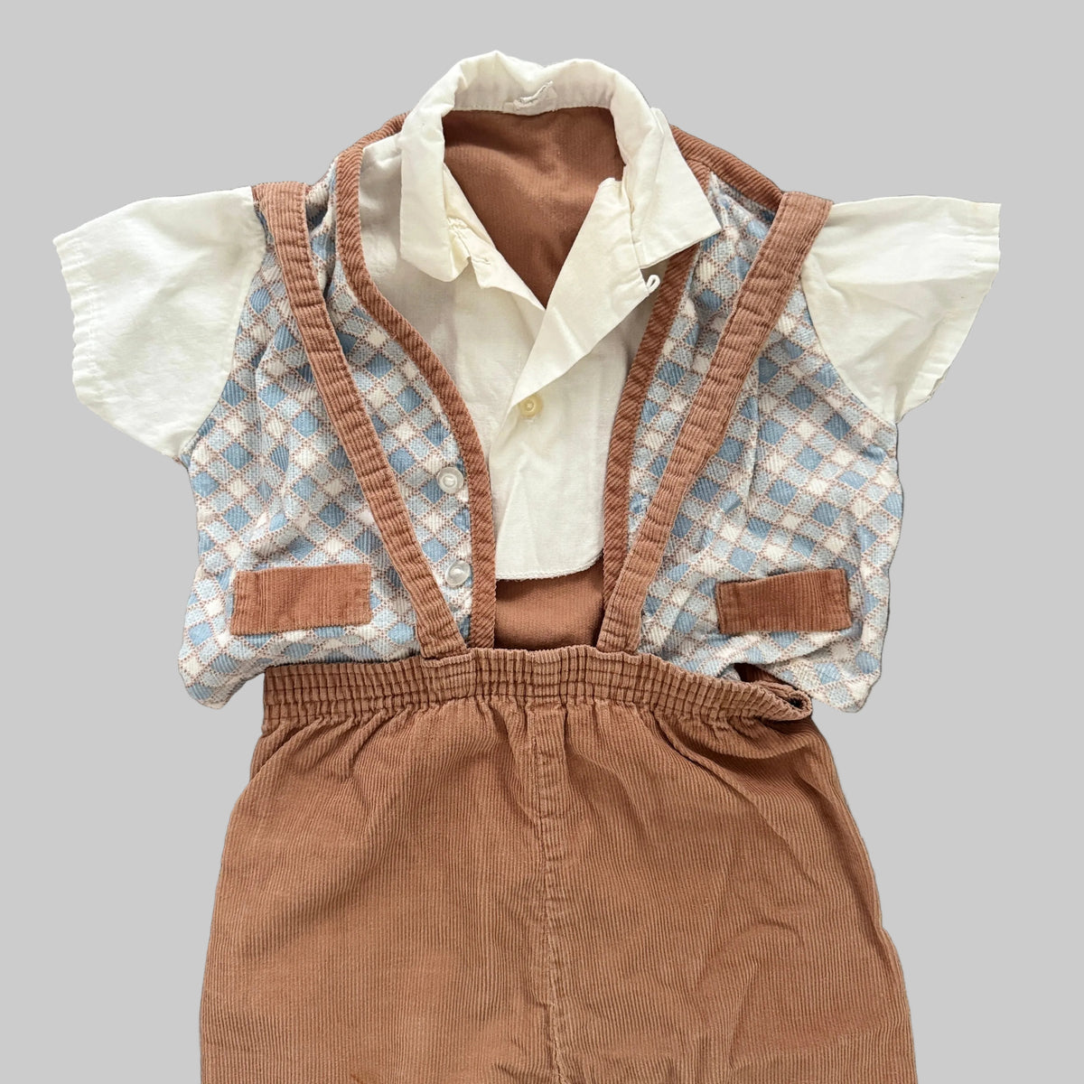 A baby boy's two-piece outfit laid flat on a textured gray fabric background. The set includes a white short-sleeve button-up shirt and a brown corduroy suspenders vest with a light blue and white argyle patterned front panel and brown trim.