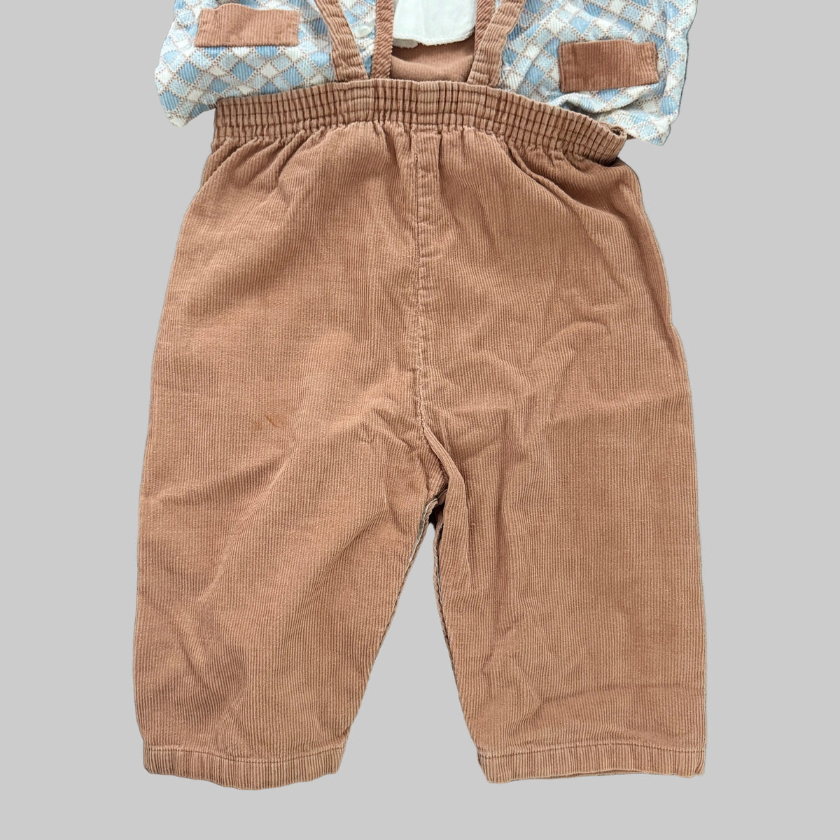 Toddler boy's corduroy overall in a warm tan-brown color, laid flat on a textured gray blanket with a light blue and white checkered shirt partially visible underneath.