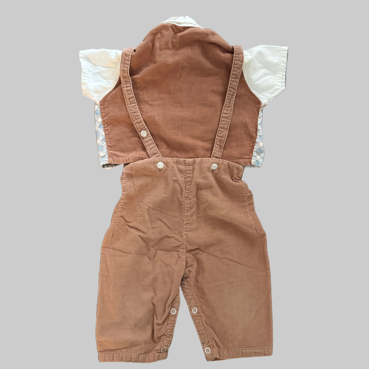 A baby's short-sleeve corduroy overall romper in a warm brownish-tan color, featuring a white long-sleeve shirt underneath with a light blue and white patterned cuff, laid flat on a textured gray fabric background.