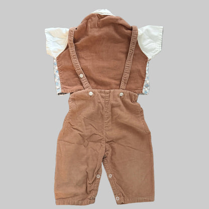 A baby's short-sleeve corduroy overall romper in a warm brownish-tan color, featuring a white long-sleeve shirt underneath with a light blue and white patterned cuff, laid flat on a textured gray fabric background.