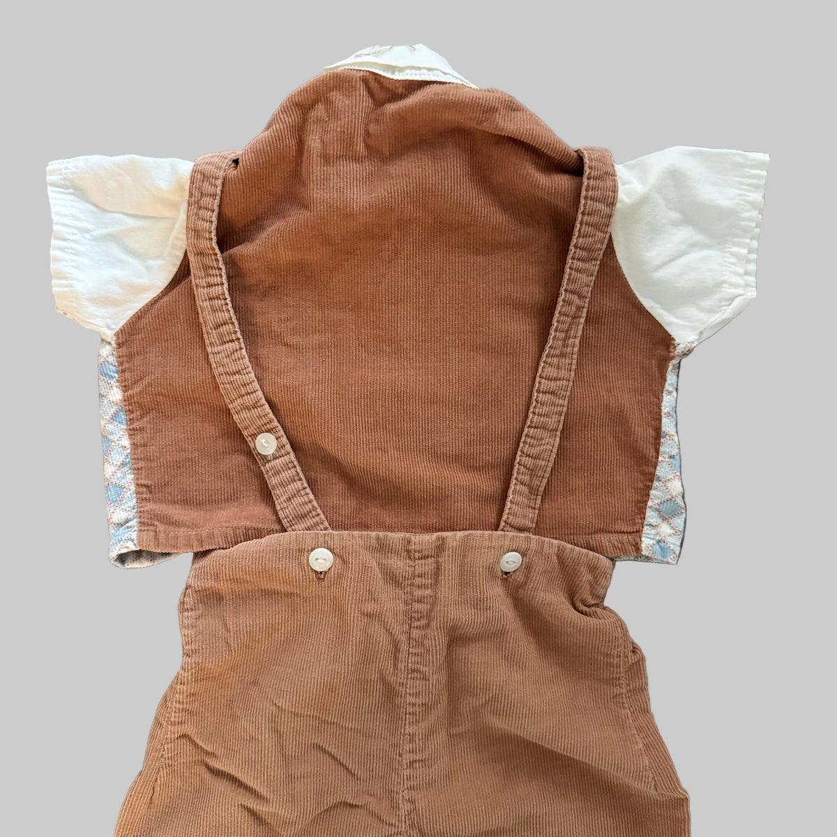 A baby's corduroy overall romper in a warm brown color, laid flat on a textured gray blanket. It features a white short-sleeve shirt underneath with patterned cuffs, secured by metal snap buttons.