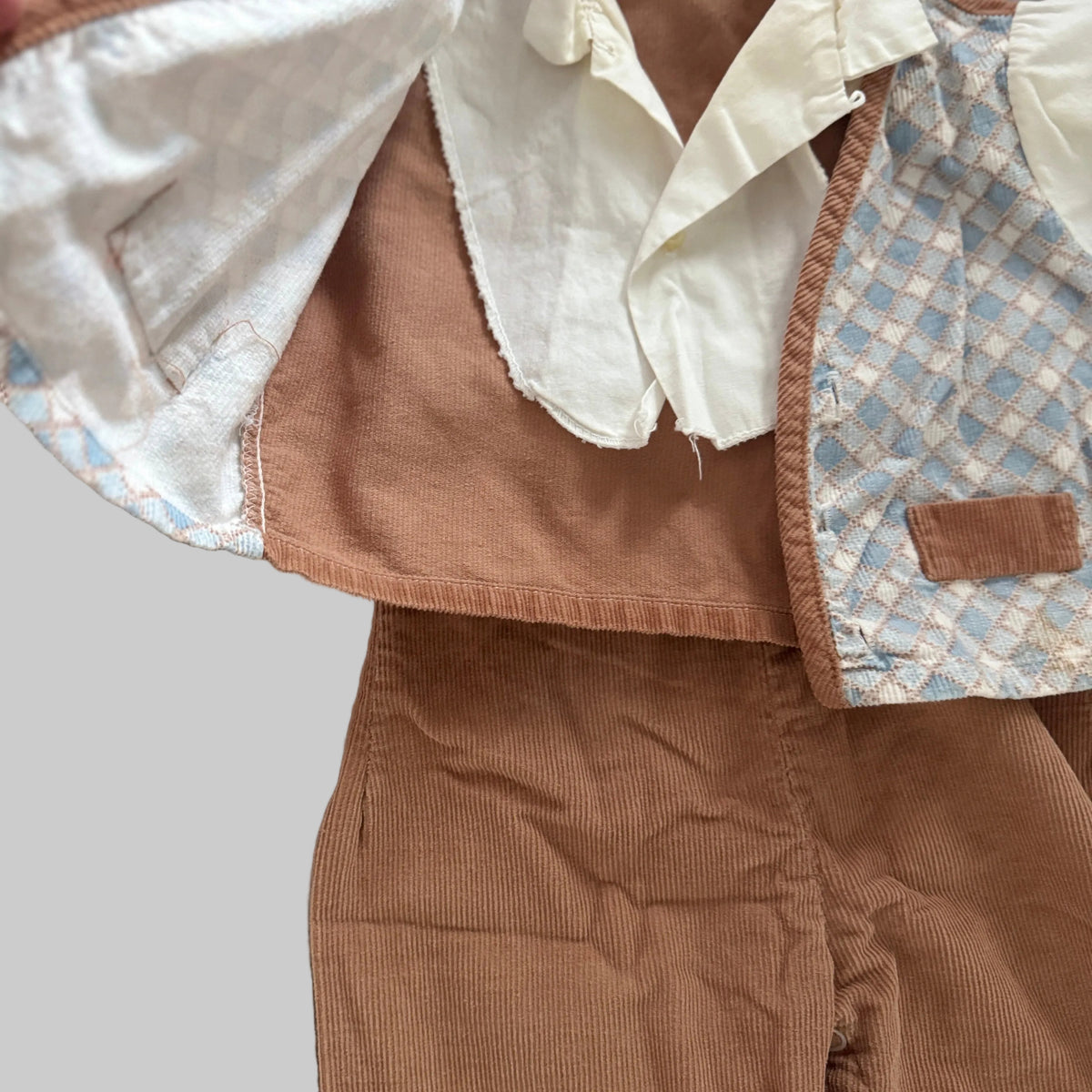 A close-up of a vintage children's clothing set featuring a brown corduroy jacket and matching trousers, layered with a cream-colored shirt and a light blue and beige patterned vest.