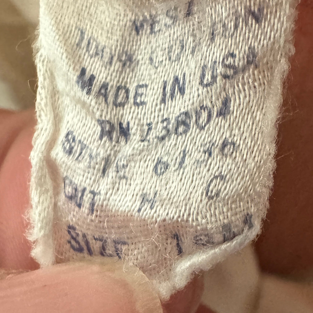 A close-up of a person's fingers holding a beige cotton garment tag with printed blue text showing 'WESTERN', '100% COTTON', 'MADE IN USA', and size information.