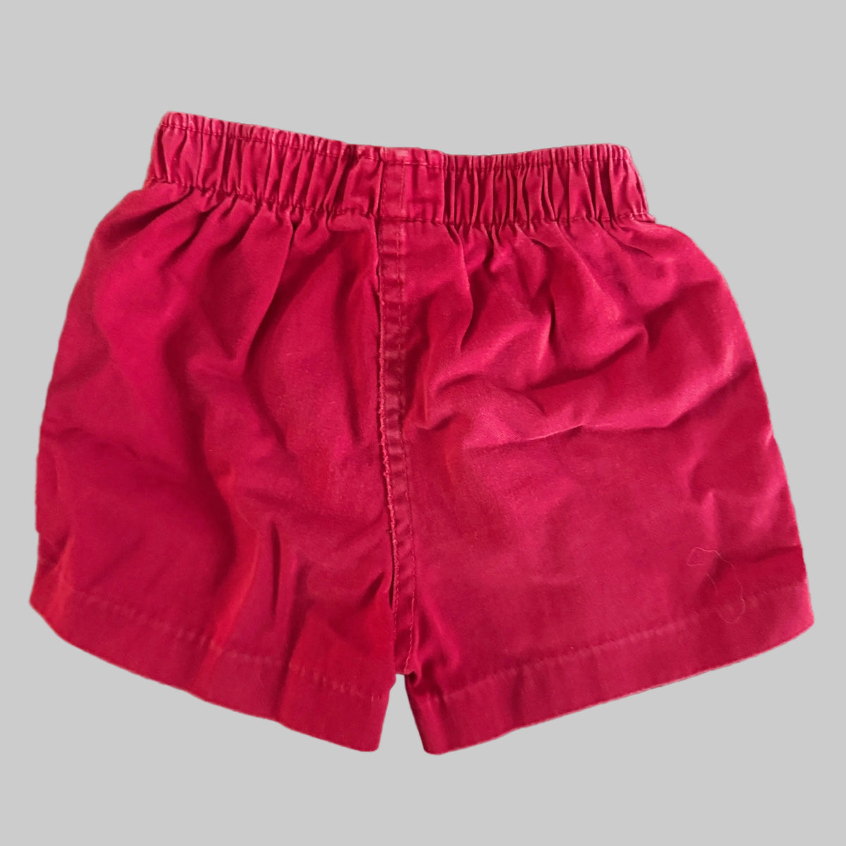 A pair of bright pinkish-red cotton shorts with an elastic waistband, laid flat on a textured beige fabric background.
