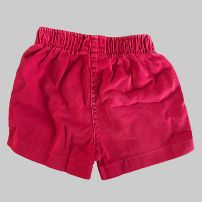 A pair of bright pinkish-red cotton shorts with an elastic waistband, laid flat on a textured beige fabric background.