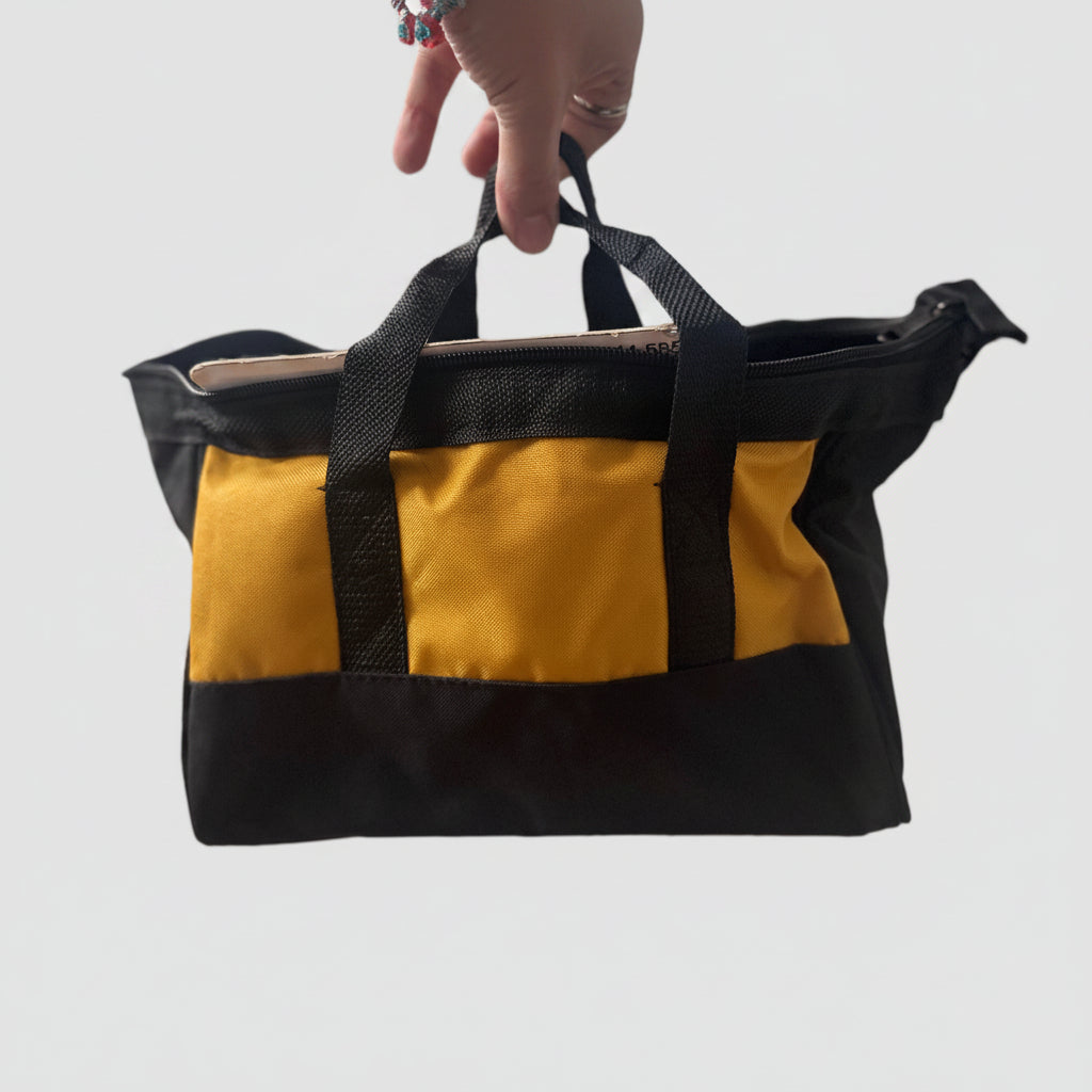 A black and mustard yellow canvas tote bag being held by a hand in an indoor setting with a blurred background.