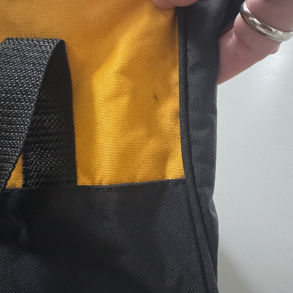 Close-up of a black and yellow fabric bag with a black webbed strap, held by a hand against a beige carpet background.