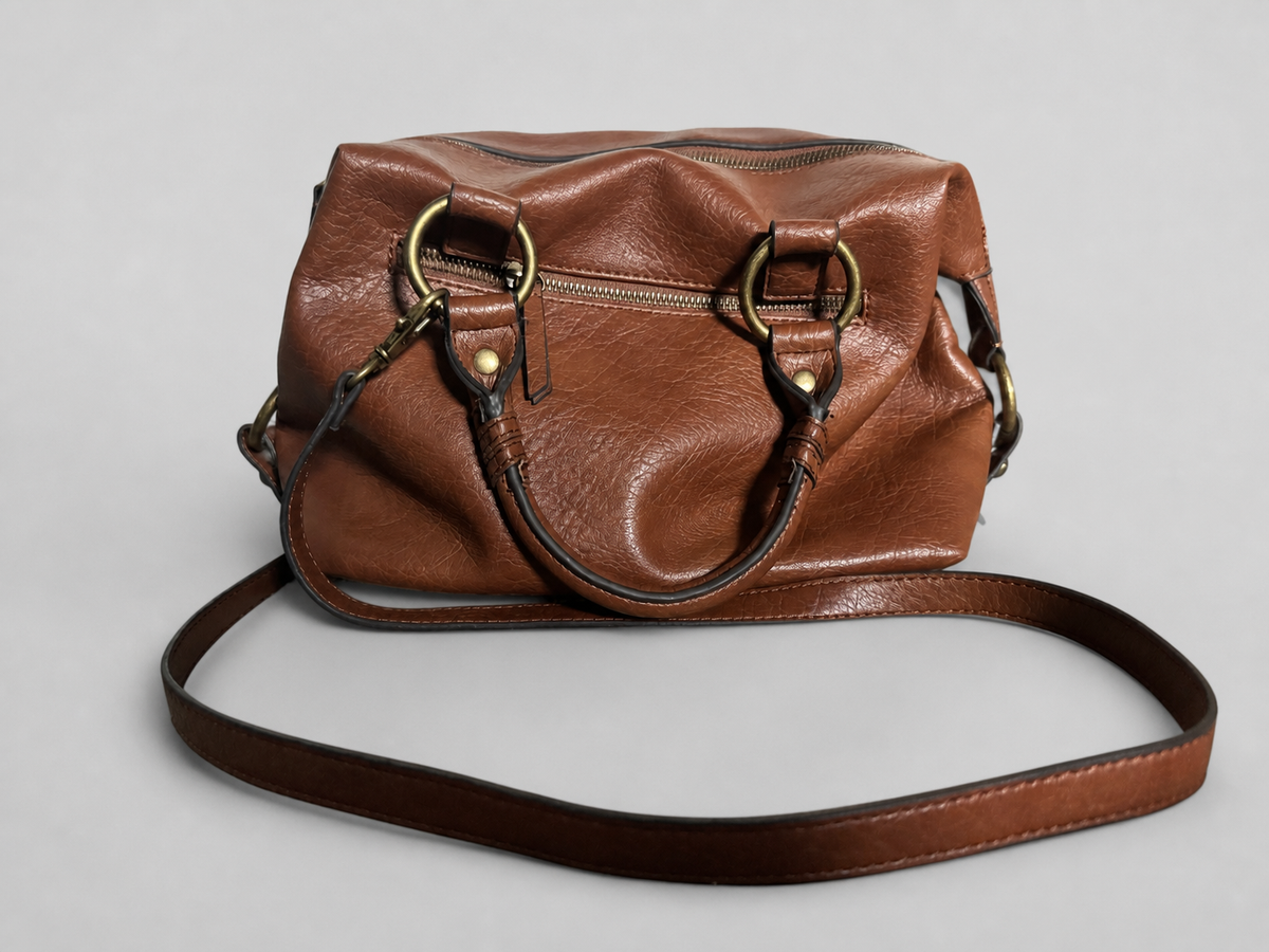 A brown leather handbag with a slouchy, rounded silhouette, featuring brass-tone hardware, a top zipper, and a detachable shoulder strap, photographed on a neutral light gray background.