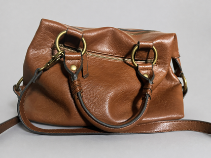 A brown leather handbag with brass-tone hardware, featuring dual top handles, a shoulder strap, and a zippered top closure, photographed against a neutral gray background.