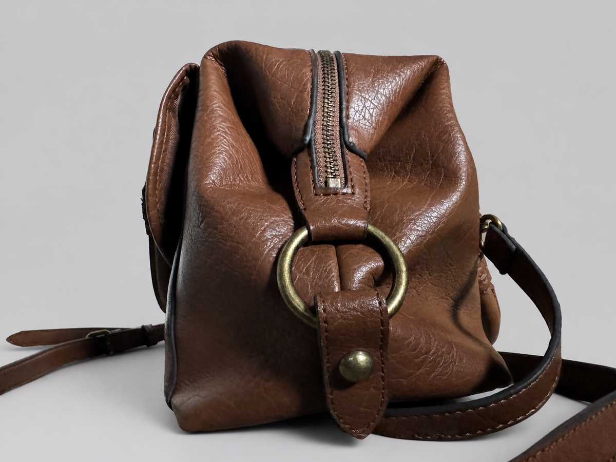 A brown leather crossbody bag with a large brass ring detail on the front, a vertical zipper, and a matching leather strap, shown against a neutral gray background.
