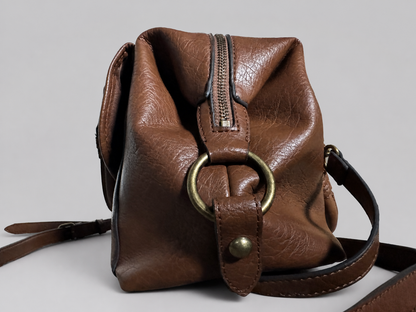 A brown leather crossbody bag with a large brass ring detail on the front, a vertical zipper, and a matching leather strap, shown against a neutral gray background.
