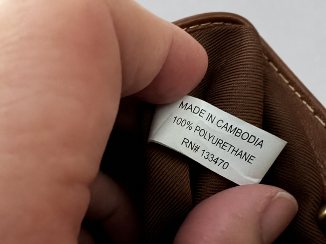 A close-up of a hand holding the opening of a brown fabric item, showing a sewn-on white label that reads 'MADE IN CAMBODIA 100% POLYURETHANE RN# 133470'.