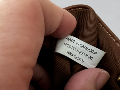 A close-up of a hand holding the opening of a brown fabric item, showing a sewn-on white label that reads 'MADE IN CAMBODIA 100% POLYURETHANE RN# 133470'.