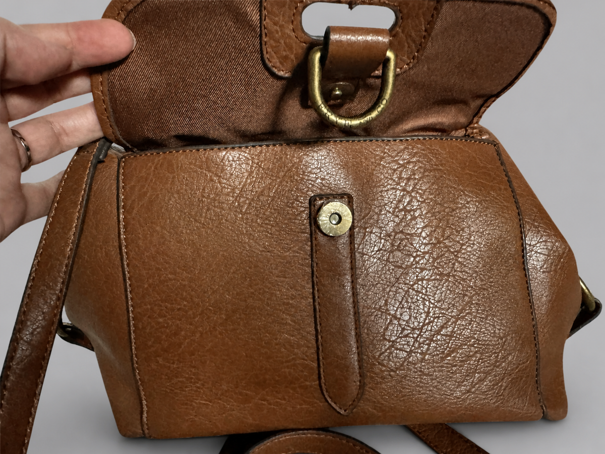 A close-up of a brown textured leather handbag being held open by a hand, showing a brass D-ring closure and a front strap detail with a snap button.