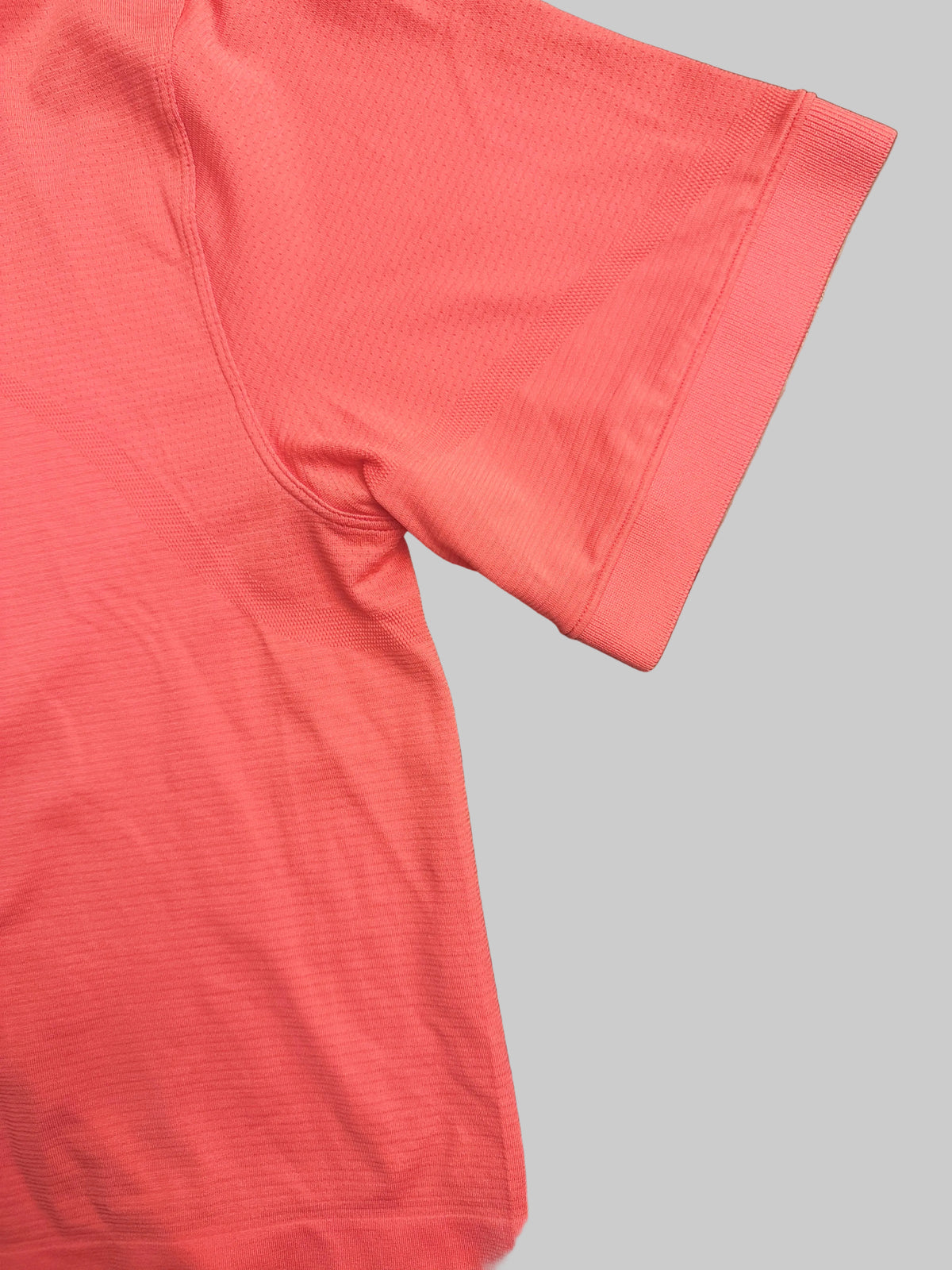 A close-up of a coral pink athletic t-shirt laid flat on a dark wooden table, showing the ribbed texture of the fabric and the sleeve cuff.
