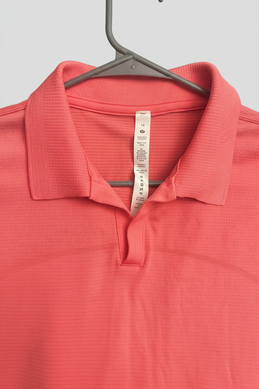 A coral pink polo shirt with a ribbed texture, hanging on a dark gray plastic hanger. The inside collar shows a white care label.
