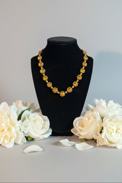 A gold-toned beaded necklace with round floral beads displayed on a black velvet jewelry bust. The necklace is photographed in a softly blurred indoor setting with a light-colored textured surface beneath the bust.