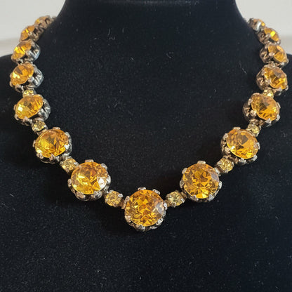 A close-up of a vintage-style necklace featuring large faceted amber-yellow rhinestones in silver-toned settings, displayed on a dark textured background.