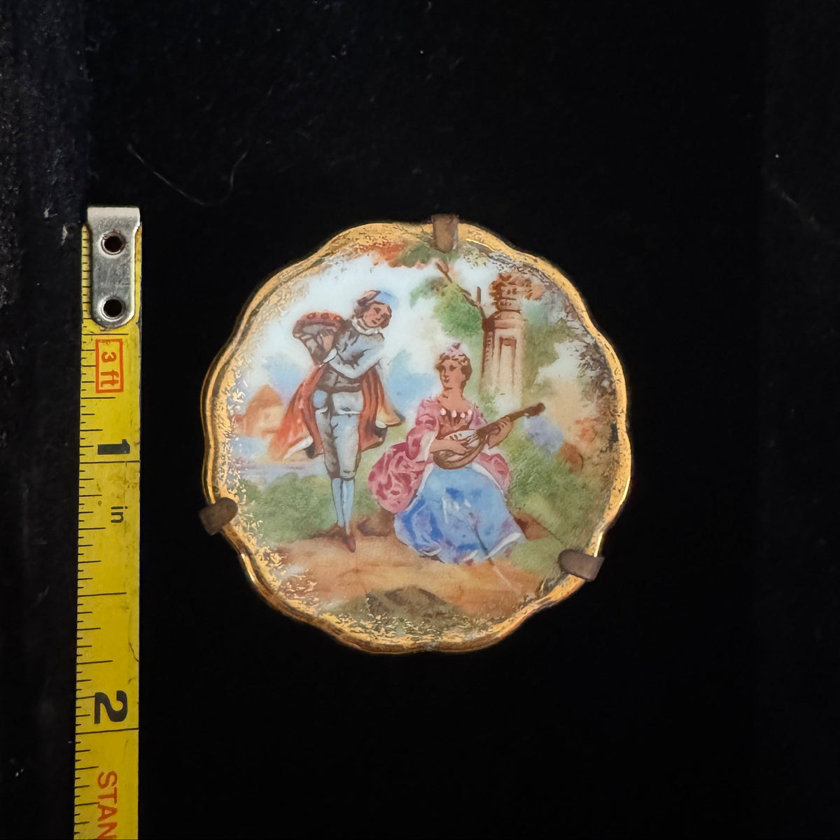 A small oval decorative plate or medallion with a hand-painted classical scene, displayed next to a yellow Stanley tape measure against a black background.