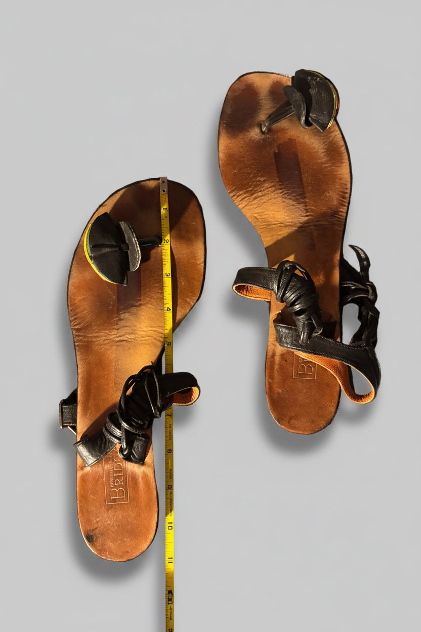 A pair of brown leather thong sandals laid flat on a textured gray fabric surface, with a yellow tape measure placed vertically between the sandals to indicate size.