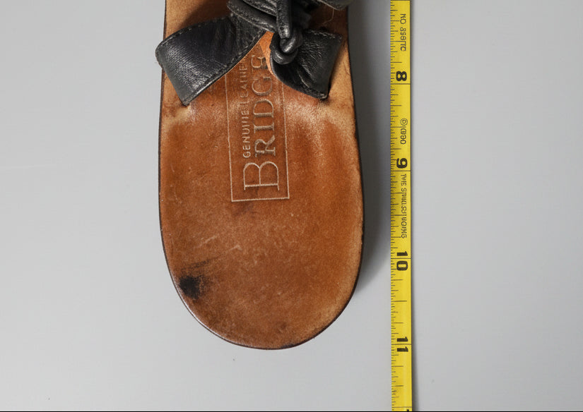 A single brown leather sandal shown from the bottom, with a black woven strap and a yellow measuring tape running vertically along the right side. The insole is embossed with the brand name 'BRIDGE' and 'GENUINE LEATHER'.