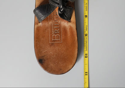 A single brown leather sandal shown from the bottom, with a black woven strap and a yellow measuring tape running vertically along the right side. The insole is embossed with the brand name 'BRIDGE' and 'GENUINE LEATHER'.