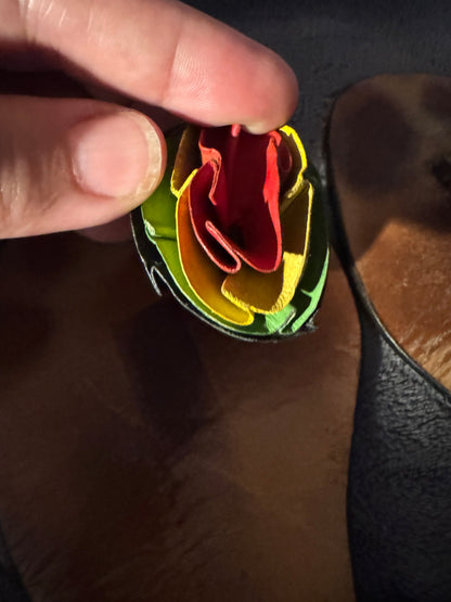A close-up of a hand holding a small, multi-layered fabric flower or rosette in red, yellow, orange, and green, set against a dark textured background.