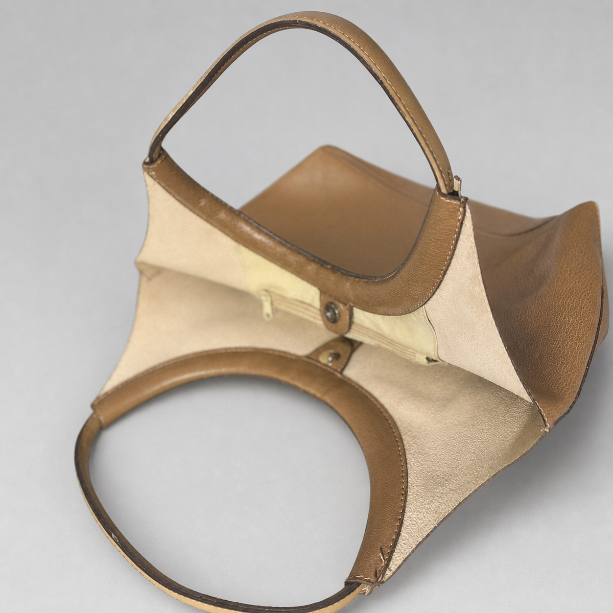 A structured leather handbag with a unique asymmetrical design, featuring a warm brown pebbled leather exterior and a light beige suede or velvet interior lining, shown on a neutral gray background.