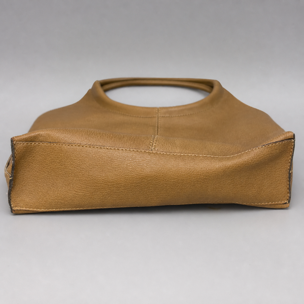 A tan leather handbag with a structured, slouchy silhouette and a single top handle, photographed against a neutral gray background.