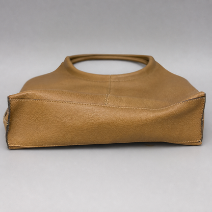 A tan leather handbag with a structured, slouchy silhouette and a single top handle, photographed against a neutral gray background.