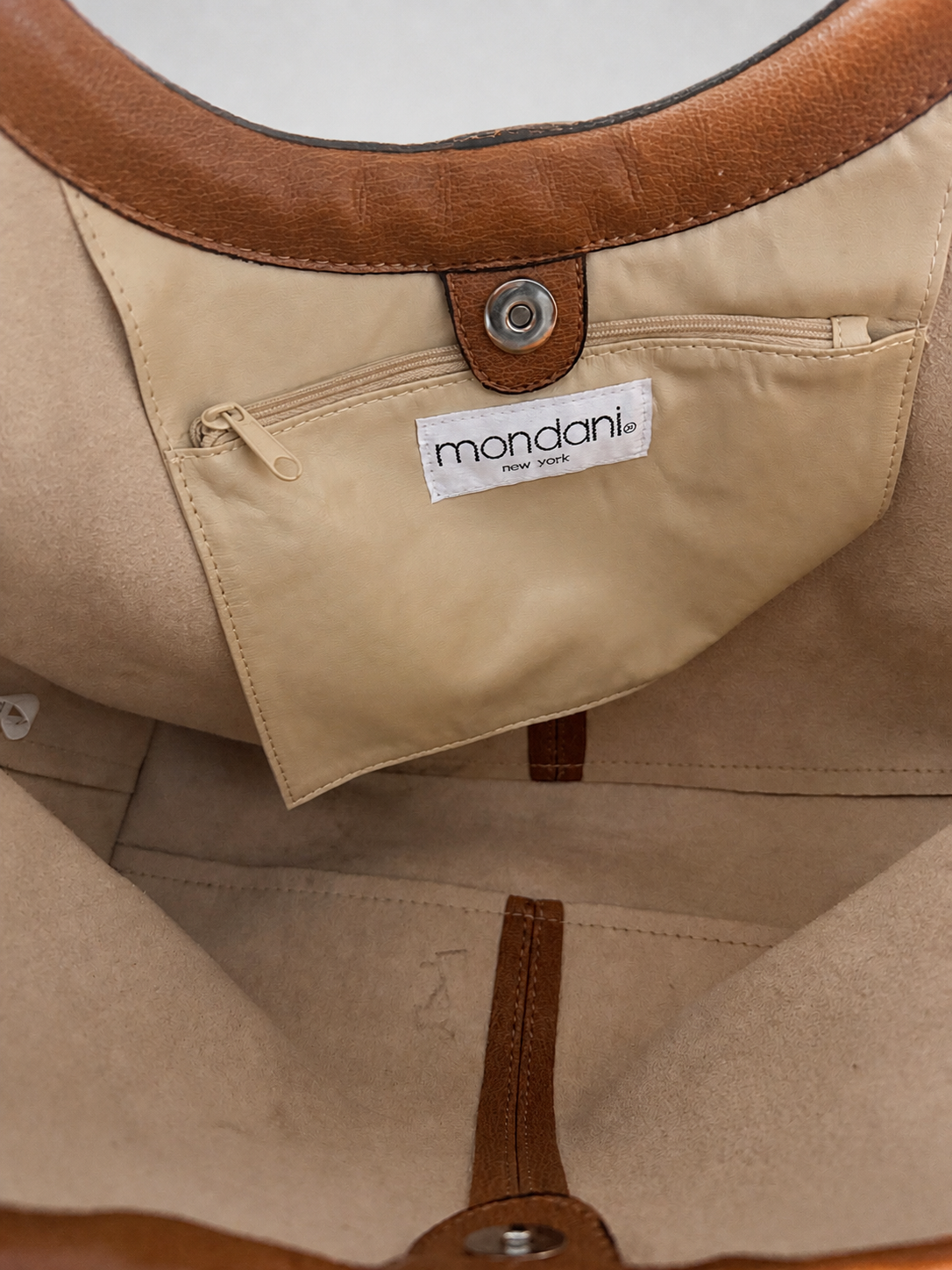 Close-up of a beige tote bag interior showing a zippered pocket with a white label reading mandarin, tan leather trim, and a metal snap closure.