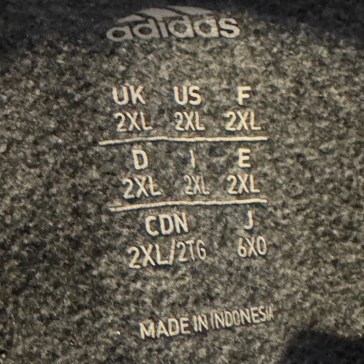 Close-up of a dark gray Adidas garment label showing size conversion information for UK, US, F, D, J, CDN, and MADE IN INDONESIA.