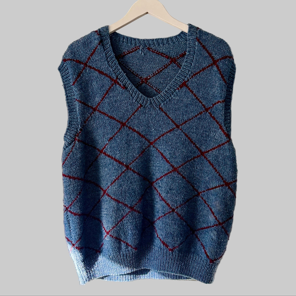 A blue sleeveless knit sweater vest with a red argyle pattern, hanging on a light wood hanger against a solid gray background.