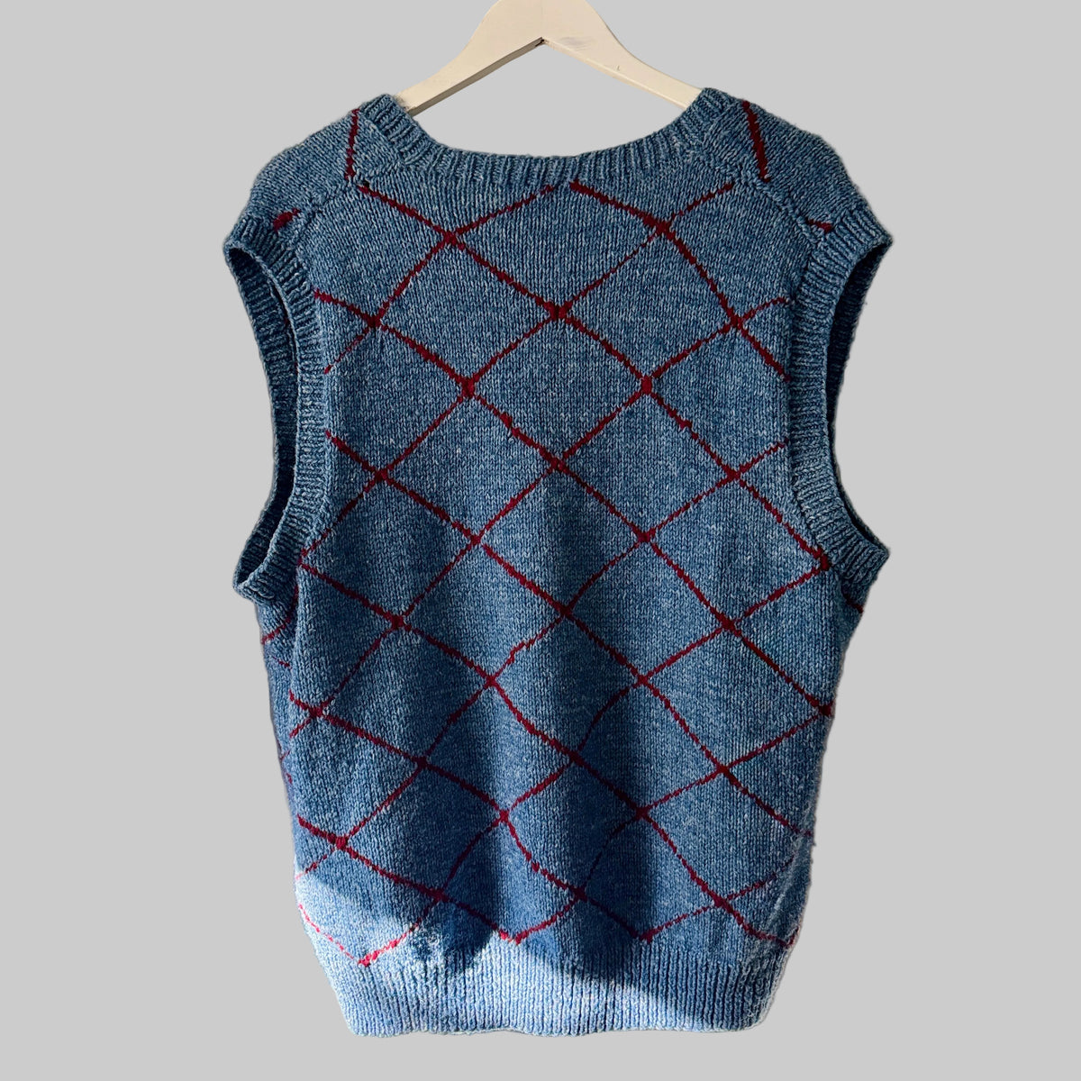 A sleeveless blue knit sweater vest with a red argyle diamond pattern, hanging on a white hanger against a white paneled door.