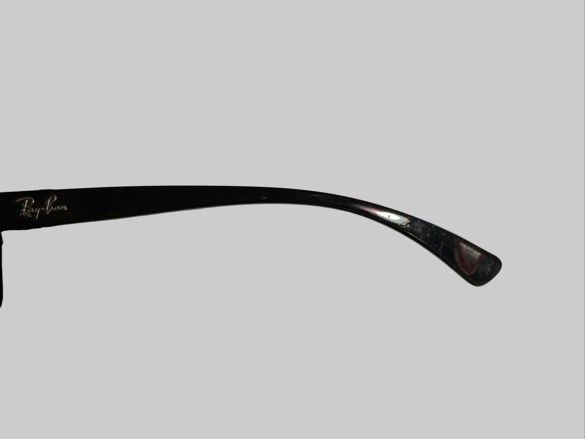 A close-up of a single black Ray-Ban sunglasses arm resting on a white textured surface, with a hand holding the other side of the frame against a dark background.