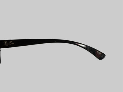 A close-up of a single black Ray-Ban sunglasses arm resting on a white textured surface, with a hand holding the other side of the frame against a dark background.