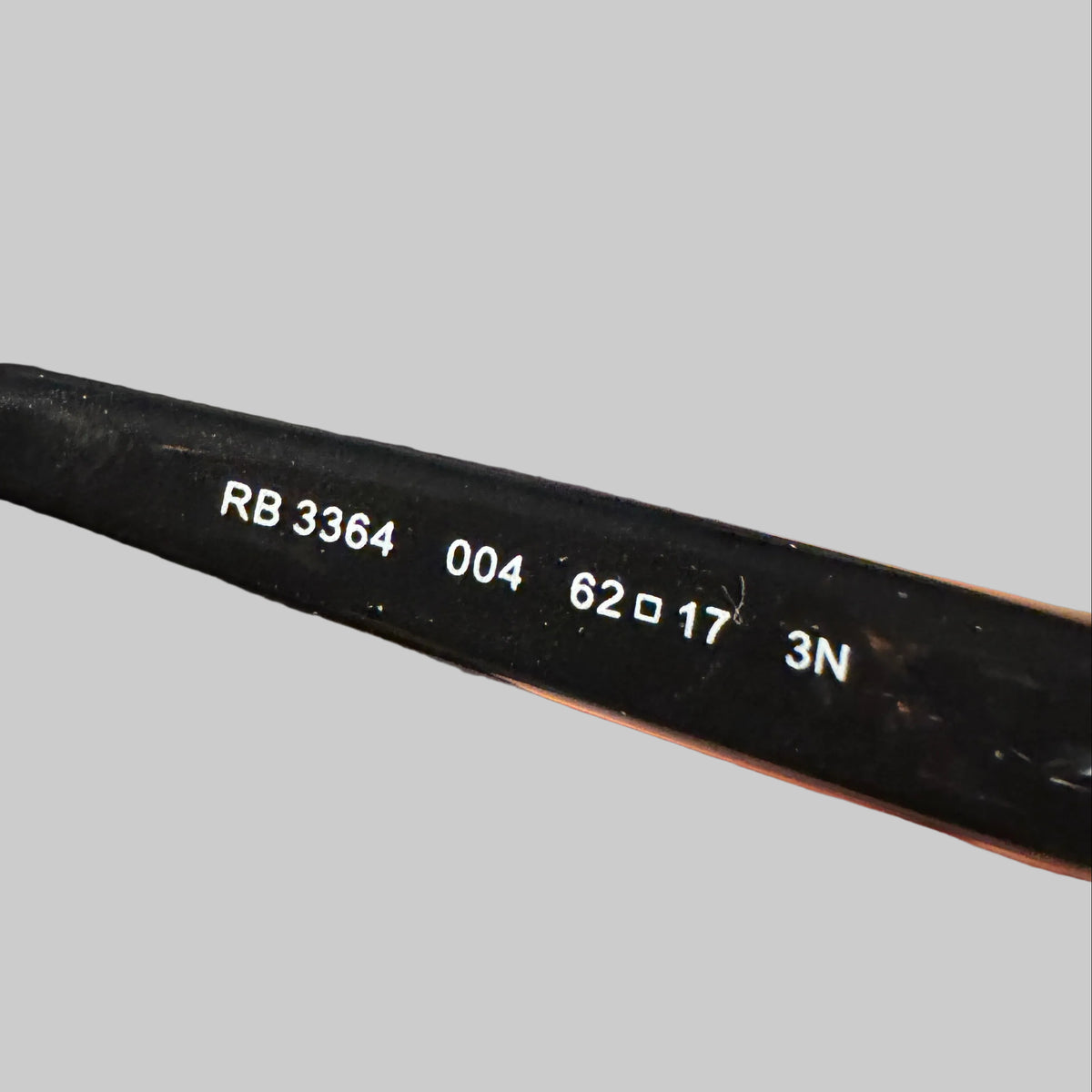Close-up of a hand holding a black eyeglass frame arm, showing white printed codes (RB 3364, 004, 62x17, 3N) on the temple.