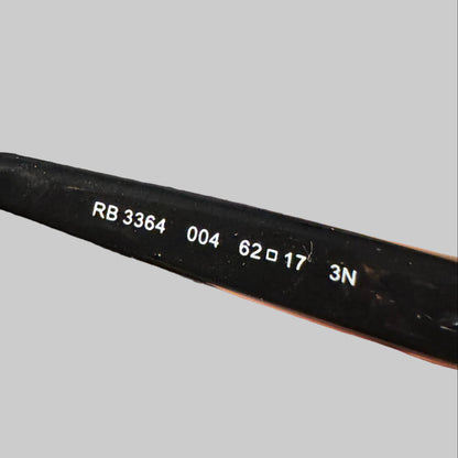 Close-up of a hand holding a black eyeglass frame arm, showing white printed codes (RB 3364, 004, 62x17, 3N) on the temple.
